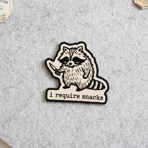 May include: Embroidered patch featuring a raccoon holding a knife. The raccoon is black and white with a fluffy body and a bandit mask. The patch has the text "i require snacks" in a black font.