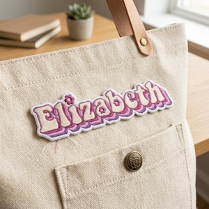 May include: A beige canvas tote bag featuring a tan leather handle attached with a gold rivet. An embroidered patch displays the name Elizabeth in a layered, retro-style font with pink and purple gradients and small flower accents.