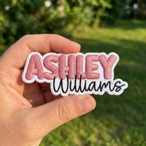 May include: A custom embroidered patch featuring the name Ashley in bold, dusty rose block letters on a white background, with the name Williams written below in a black cursive script font, all outlined with a white border.