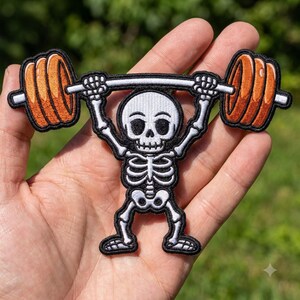 May include: An embroidered patch featuring a white skeleton with black outlines lifting a barbell with bright orange weight plates. The skeleton has a round skull, visible ribcage, and is shown in a standing position while holding the bar overhead.