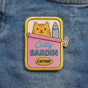 May include: Embroidered patch featuring a cartoon cat in a sardine can. The patch has a yellow border, a pink can, and the words "Catty Sardin" in teal. A fish and the word "Catnip" are also visible.
