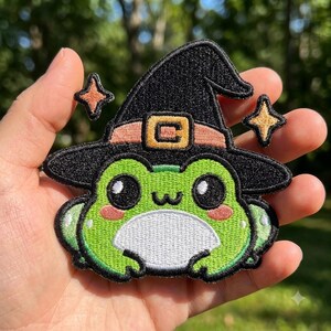 May include: An embroidered patch featuring a bright green frog with large black eyes, pink cheeks, and a white belly. The frog wears a black witch hat with a peach-colored band and a gold buckle, flanked by two small, four-pointed gold stars.