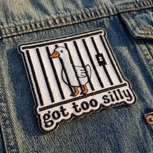 May include: Embroidered patch featuring a white goose inside a cage with black bars. The goose has an orange beak and feet. The patch is outlined in black and has the words "got too silly" in a playful font.