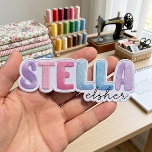 May include: An embroidered patch featuring the name STELLA in large, puffy, pastel-colored block letters, with the name elsher written in black cursive script underneath. The patch has a white border and is held against a sewing-themed background.