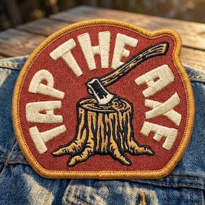 May include: A circular embroidered patch with a red background and a gold border. The patch features a tree stump with an axe embedded in the top, surrounded by the white embroidered text "TAP THE AXE" in a bold, sans-serif font style.