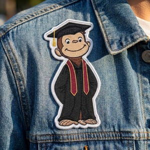 May include: An embroidered patch featuring a brown monkey wearing a black graduation cap with a gold tassel and a black graduation gown with a maroon stole. The patch is attached to a blue denim jacket, showcasing detailed stitching and outlines.