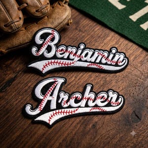 May include: Two embroidered patches featuring the names Benjamin and Archer in a white script font with black outlines. Each name is underlined by a white swoosh detailed with red baseball-style stitching patterns on a black background.