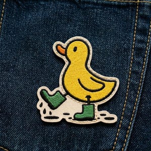 May include: Embroidered patch featuring a yellow duck wearing green boots, with a black outline. The duck is standing in a puddle of green water. The patch is on a blue denim background.