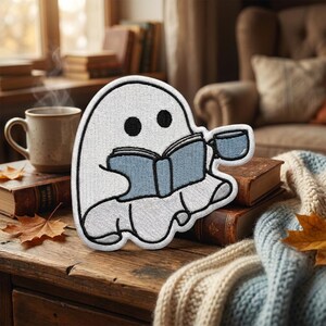May include: An embroidered patch shaped like a white ghost with two black circular eyes. The ghost is holding an open blue book in its hands and a small blue mug in one hand. The patch features a thick black outline and detailed stitching.