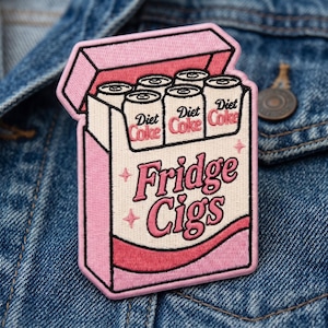 May include: An embroidered patch shaped like a cigarette pack in shades of pink and cream. The design features six soda cans labeled Diet Coke at the top and the text Fridge Cigs in pink cursive lettering across the center with small star accents.