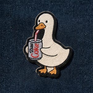 May include: Embroidered patch featuring a white goose with an orange beak and feet, holding a Diet Coke can with a red straw. The patch has a black border and is set against a dark blue denim background.