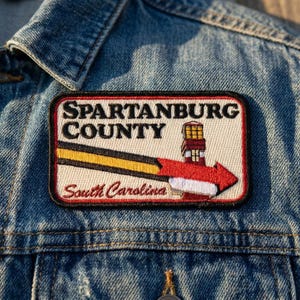 May include: A rectangular embroidered patch featuring the black text SPARTANBURG COUNTY and South Carolina in red script. The design includes a yellow and black diagonal stripe, a red arrow, and a lighthouse graphic on a cream background.