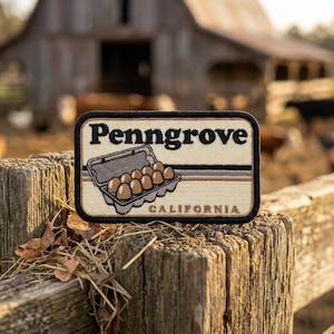 May include: A rectangular embroidered patch with a cream background and a black border. It features the text "Penngrove" in black, an illustration of a carton of brown eggs, and the word "CALIFORNIA" in brown, with horizontal stripes.