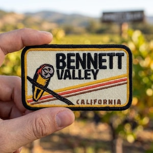 May include: A rectangular embroidered patch with a cream background and a black border. It features a stylized owl illustration, the text BENNETT VALLEY in black, and CALIFORNIA in brown, with diagonal orange and brown stripes across the center.