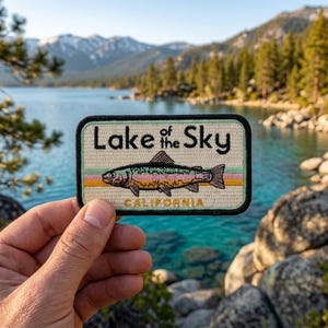 May include: A rectangular embroidered patch with a black border, featuring the text Lake of the Sky in black lettering above a central illustration of a rainbow trout. The word CALIFORNIA is printed in yellow below the fish on a cream background.