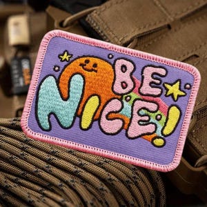 May include: A rectangular patch with the words "BE NICE!" in colorful bubble letters. The patch features a purple background with a pink border and an orange cartoon character. A yellow star and other small details are included.