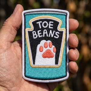 May include: A rectangular embroidered patch featuring a teal background with a black shield-shaped emblem. The text "TOE BEANS" is stitched in white above a white cat paw graphic with orange pads, all outlined in white, black, and gold thread.