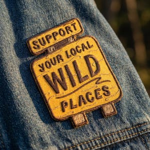 May include: A rectangular embroidered patch with a yellow background and dark brown stitched text that reads SUPPORT YOUR LOCAL WILD PLACES. The patch is shaped like a signpost with two small posts at the bottom, attached to blue denim fabric.