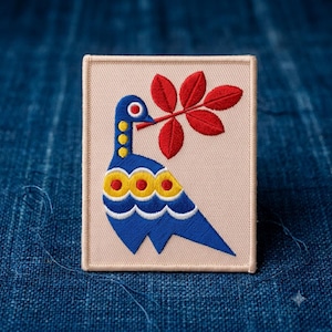 May include: A rectangular embroidered patch on a light beige background featuring a stylized blue bird with yellow and red circular accents. The bird holds a branch with five vibrant red leaves in its beak, rendered in detailed thread work.