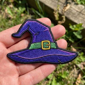 May include: An embroidered patch shaped like a pointed witch hat. The hat is deep purple with a wide brim and a folded top. It features a bright green band around the base with a prominent yellow buckle, all outlined in thick black stitching.