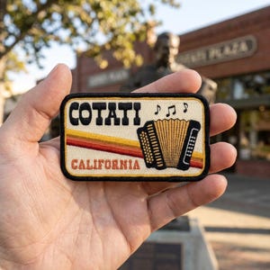 May include: A rectangular embroidered patch with a cream background, featuring the black text COTATI and CALIFORNIA. It displays a black accordion, three black musical notes, and three horizontal stripes in yellow, orange, and red.