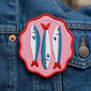May include: Embroidered patch featuring three stylized fish with blue and white bodies, red tails, and black eyes, set against a pink background. The patch has a scalloped red border and is attached to a denim jacket.