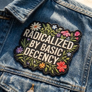 May include: A black embroidered patch with a scalloped edge featuring the white text RADICALIZED BY BASIC DECENCY. The design is surrounded by colorful embroidered flowers and green leaves, attached to a blue denim fabric background.