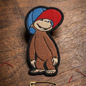 May include: Embroidered patch featuring a cartoon monkey wearing a red and blue hat. The monkey is brown with a black outline. The patch is on a wooden surface.