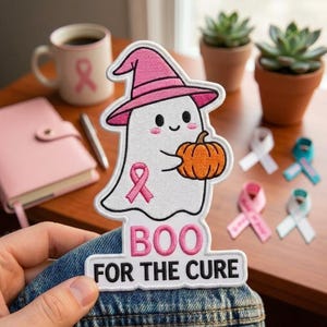 May include: An embroidered patch featuring a white ghost wearing a pink witch hat and holding an orange pumpkin. The ghost has a pink breast cancer awareness ribbon on its body. Below the ghost, the text reads BOO FOR THE CURE in pink and black.