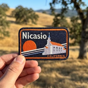 May include: A rectangular embroidered patch with a dark blue background and an orange border. It features the white text Nicasio above an orange sun, a white church building with a steeple, and the word CALIFORNIA in orange at the bottom.