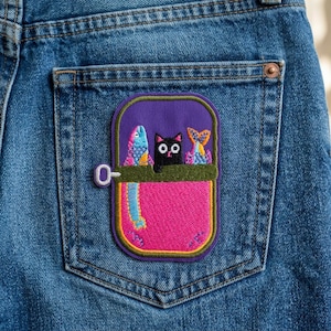 May include: Embroidered patch featuring a black cat and two colorful fish inside a sardine can design. The patch is in shades of purple, pink, green, and yellow, and is attached to the back pocket of a pair of blue denim jeans.