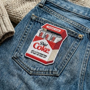 May include: Embroidered patch in the shape of a Diet Coke cigarette pack, with the text "Diet Coke" and "JUST A LITTLE SOMETHING TO TAKE THE EDGE OFF." The patch is red and white, with the words "FRIDGE CIGS" at the top. The patch is sewn onto a blue denim jean pocket.
