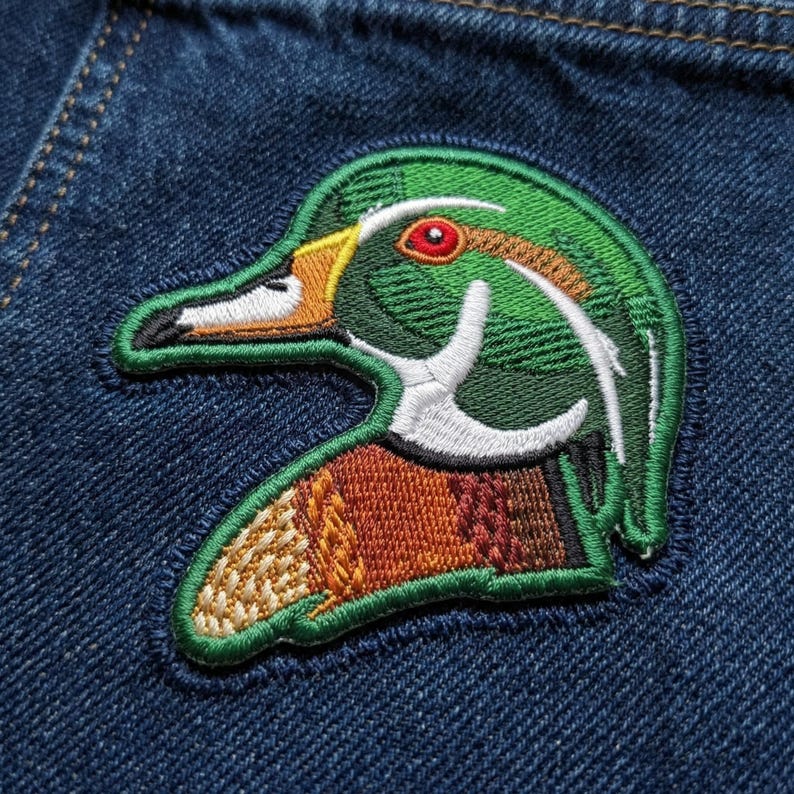 May include: Embroidered patch featuring a detailed duck head with green, brown, white, and yellow accents. The patch is set against a blue denim background, showcasing the intricate stitching and design.