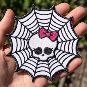 May include: An embroidered patch featuring a white skull with large black eye sockets and eyelashes, adorned with a bright pink bow. The skull is centered within a black and white spider web design that has a scalloped, pointed outer edge.