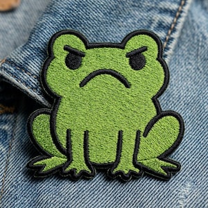 May include: Embroidered patch featuring a green frog with a grumpy expression. The frog has black outlines and details, including the eyes and mouth. The patch is set against a blue denim background.