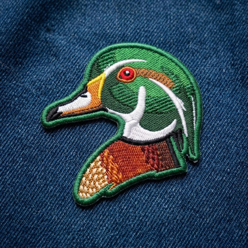 May include: Embroidered patch featuring a detailed duck head. The patch showcases a green head with white and brown accents, a yellow beak, and a red eye. The duck's chest is a mix of brown and tan. The patch is outlined in green.