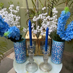 Puede incluir: Una composición decorativa con velas azules en candelabros de cristal transparente, flanqueada por jarrones florales azules y blancos. Los jarrones y candelabros están sobre una mesa redonda, con faroles dorados y flores al fondo.