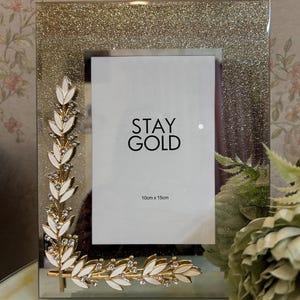 May include: A rectangular photo frame with a glittery gold border and a white mat. The frame is decorated with gold and white leaf accents and the text "STAY GOLD". The frame dimensions are 10cm x 15cm.