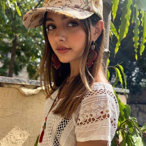Puede incluir: Una gorra de béisbol beige con un estampado floral amarillo y marrón, combinada con una blusa blanca de manga corta con intrincados detalles de encaje y bordados, junto con largos pendientes colgantes rojos y dorados y un collar de cuentas con borla roja.