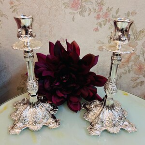 May include: Two ornate silver candlesticks with detailed floral designs. Each candlestick has a wide base, a slender stem, and a cup-shaped top. A dark red flower is placed between the candlesticks. The background features a floral wallpaper.