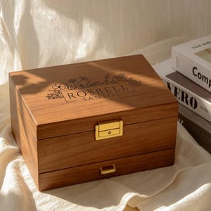 May include: A rectangular wooden jewellery box with a medium brown finish, featuring a gold-toned metal latch and a pull handle on the bottom drawer. The lid is engraved with a floral design and the text ROSBELL SANTOS in a centred rectangular frame.