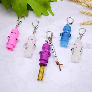 May include: A collection of colorful syringe-shaped keychains. The keychains are in pink, white, blue, and purple, each with a smiling face and a silver keyring. One keychain has a beaded tag that says "UDMFGVES".