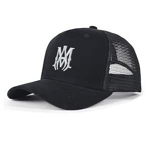 May include: A black trucker cap featuring a structured front panel with a white embroidered monogram logo consisting of the letters MA. The cap includes a curved peak and a breathable black mesh back panel with an adjustable snapback closure.