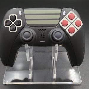 May include: A black video game controller with a unique design, featuring a D-pad and buttons in contrasting colors. The controller is displayed on a clear acrylic stand, showcasing its sleek form and design. The controller has a gray striped design.