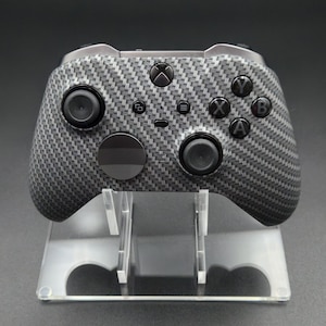 Puede incluir: Un mando de Xbox negro y gris con un patrón de fibra de carbono. El mando tiene botones y joysticks negros. Se muestra en un soporte de acrílico transparente.