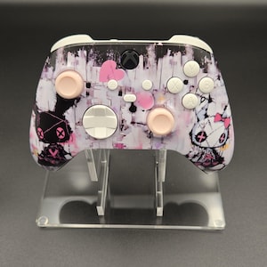 Może przedstawiać: Kontroler Xbox z niestandardowym wzorem. Posiada czarno-biały abstrakcyjny wzór z różowymi akcentami i grafiką z kreskówkowymi króliczkami. Kontroler ma białe przyciski i różowe gałki, na przezroczystym stojaku.