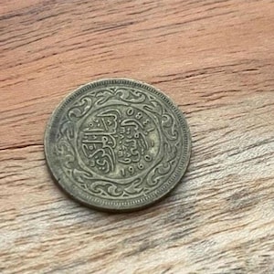 Puede incluir: Una moneda de color bronce con intrincados diseños en relieve y escritura árabe. La superficie de la moneda muestra signos de desgaste y la fecha "1980" es visible. La moneda está sobre un fondo de madera.