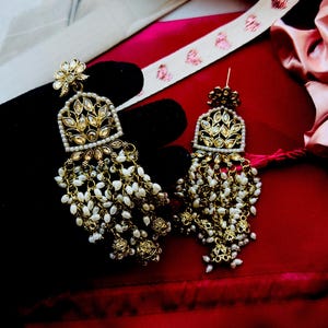 May include: A pair of gold-toned chandelier earrings with intricate leaf and floral designs. The earrings feature a cascade of small, white pearls and are set against a dark background, highlighting their ornate details. The earrings are a statement piece.
