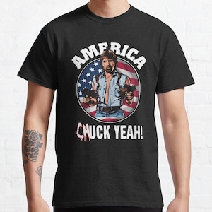 Könnte beinhalten: Schwarzes T-Shirt mit einer Grafik eines Mannes, der zwei Pistolen hält, vor einem Hintergrund der amerikanischen Flagge. Das Wort "AMERICA" steht über dem Bild, darunter "CHUCK YEAH!".