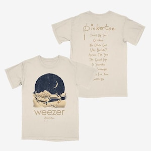 Puede incluir: Una camiseta beige de manga corta con un gráfico circular azul oscuro de un paisaje nocturno con una luna creciente y montañas en el frente. Las palabras "weezer" y "Pinkerton" están impresas debajo. La parte trasera lista títulos de canciones.
